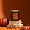 Candle labeled 'Caramel Pumpkin Spice' with pumpkins and cinnamon sticks on a stone surface against an orange background